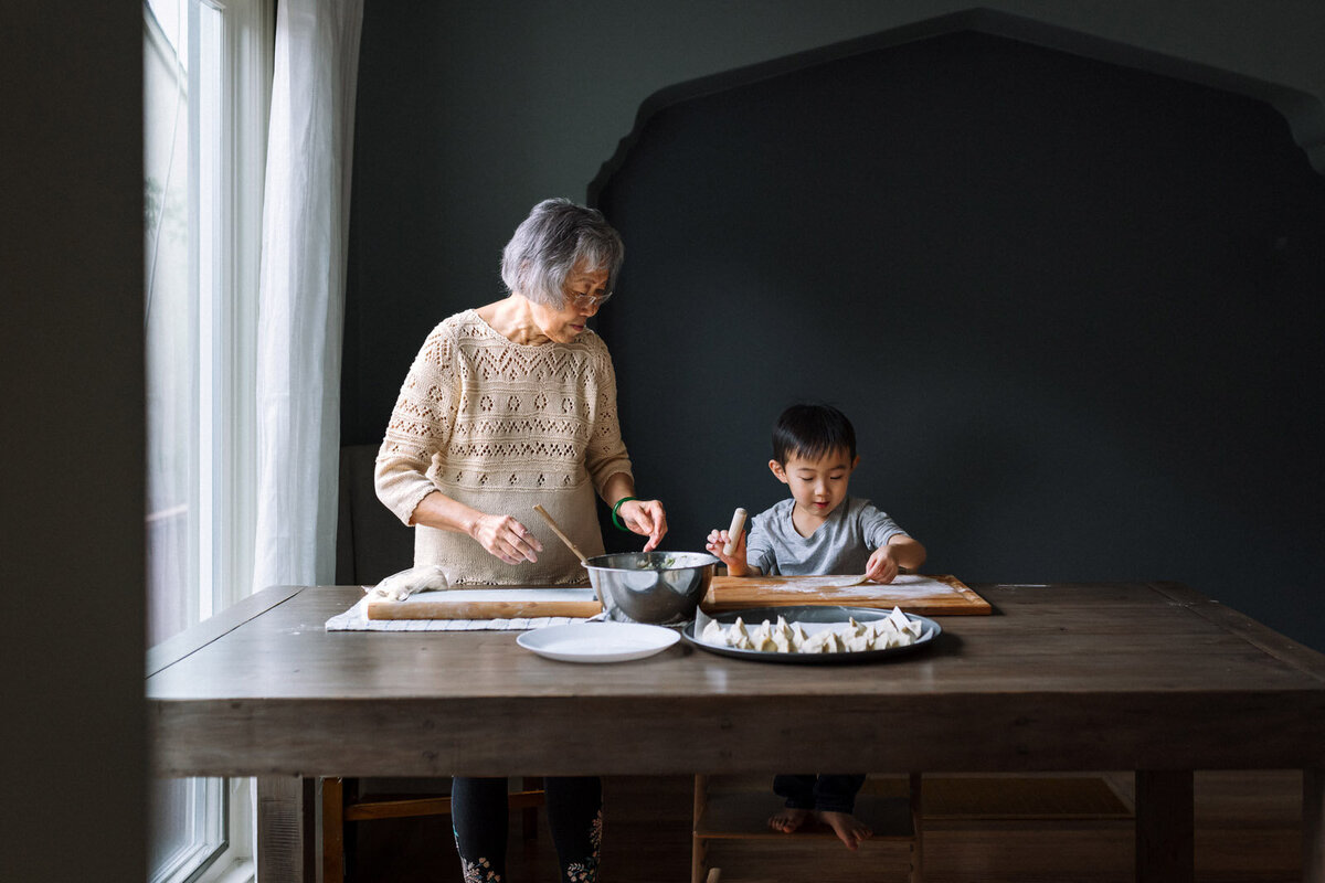 grandmother and grandson making chinese dumplings together while next to a window