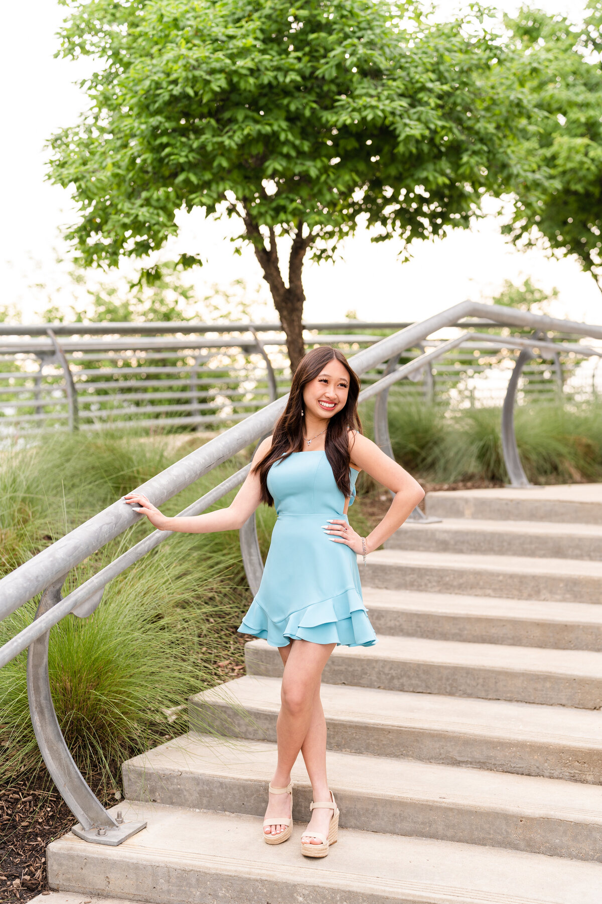 Houston high school senior girl wearing baby blue dress with hand on hip and other on a stair railing while laughing away in Buffalo Bayou Park in Downtown Houston