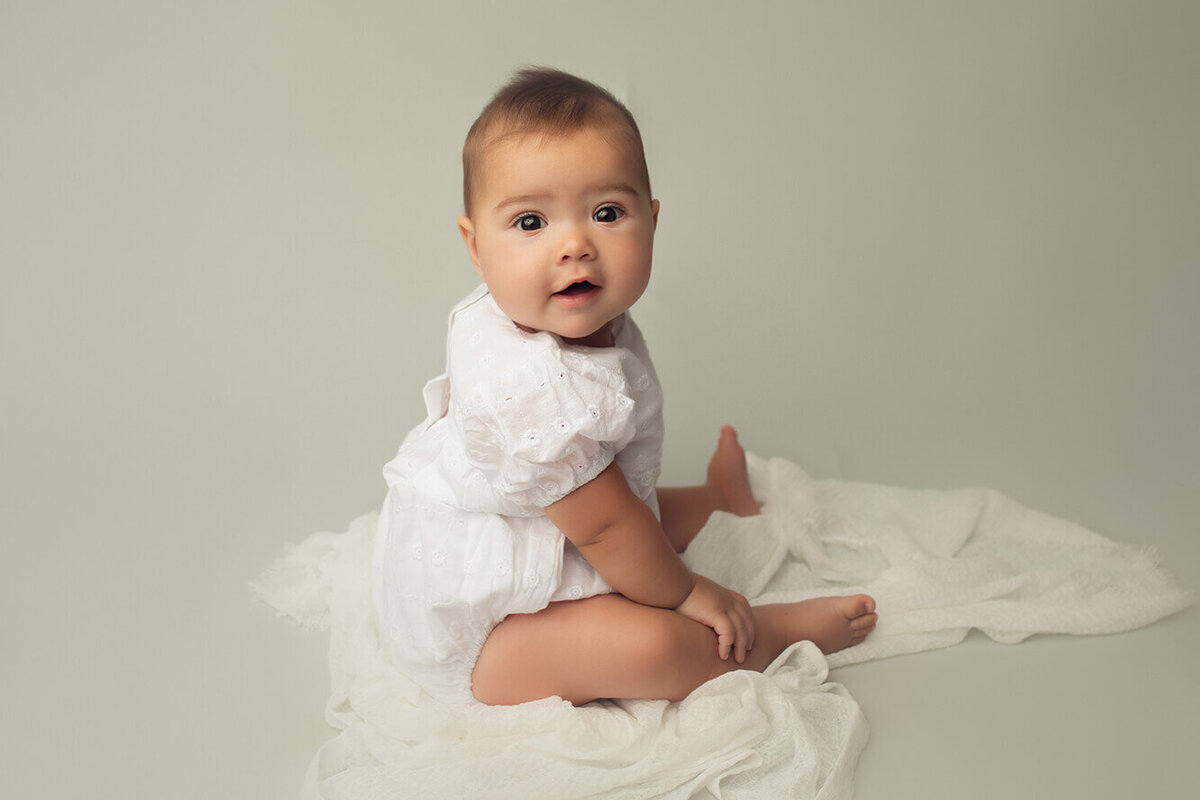 a six month old girl sitting by herself wearing a white romper photographed by lulu belle photography in rochester ny