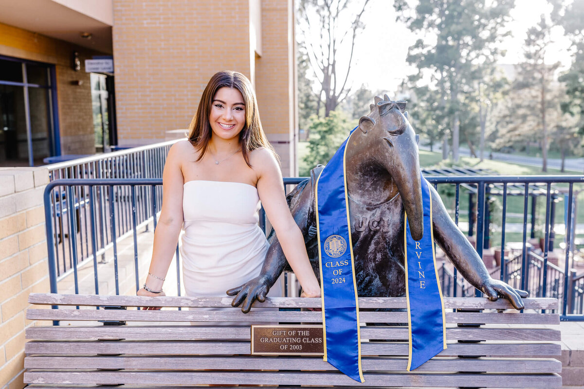A woman in a white dress stands next to a seated anteater statue wearing a blue graduation stole. The statue is on a bench with trees and a building in the background. A plaque reads For the Graduating Class of 2005.