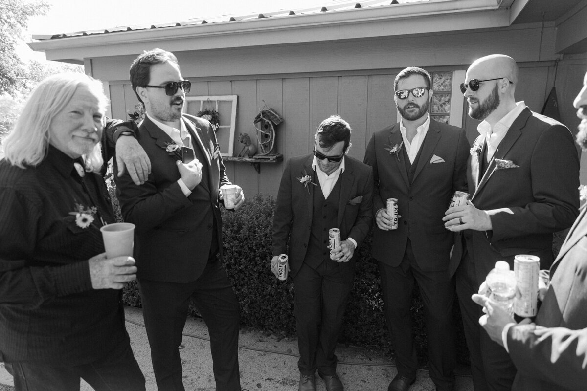 A black and white candid moment of multiple groomsmen hanging out before a wedding in Fort Worth, Texas. They all are wearing suits with boutonnieres, and many of them are holding drinks and wearing sunglasses.