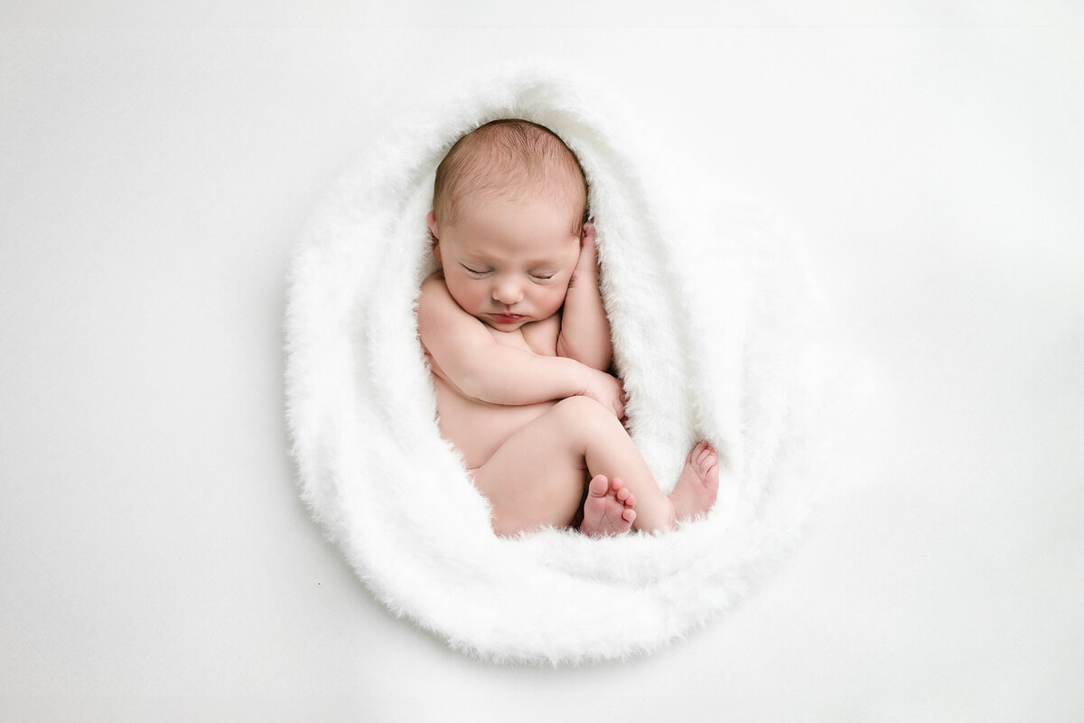 Newborn girl is wrapped in a loose fuzzy white wrap and all curled up sleeping sweetly during her neutral newborn photo session in Pittsburgh.
