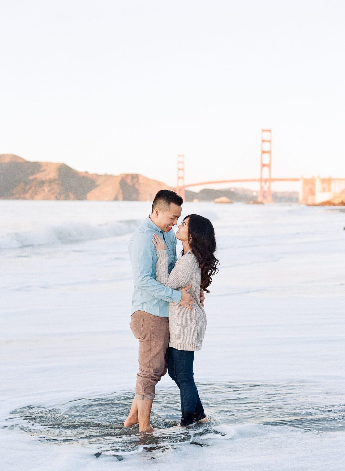 Loving couple look into each others' eyes in ankle-deep water as warm sun hues and the Golden Gate Bridge get blurred in the background.