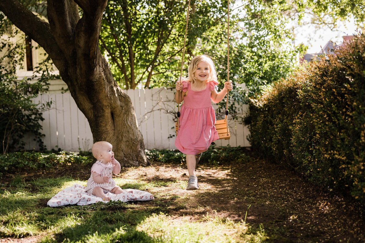 A smiling young girl in a pink dress runs through a sunlit garden toward a sitting baby, captured beautifully by a family photographer in Pittsburgh, PA.