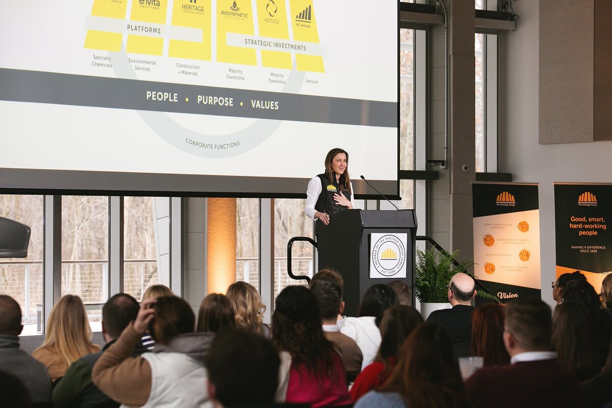 a-professional-speaker-stands-at-the-podium-addressing-an-audience-at-the-heritage-group-conference-in-indianapolis-with-a-presentation-slide-displayed-behind-her