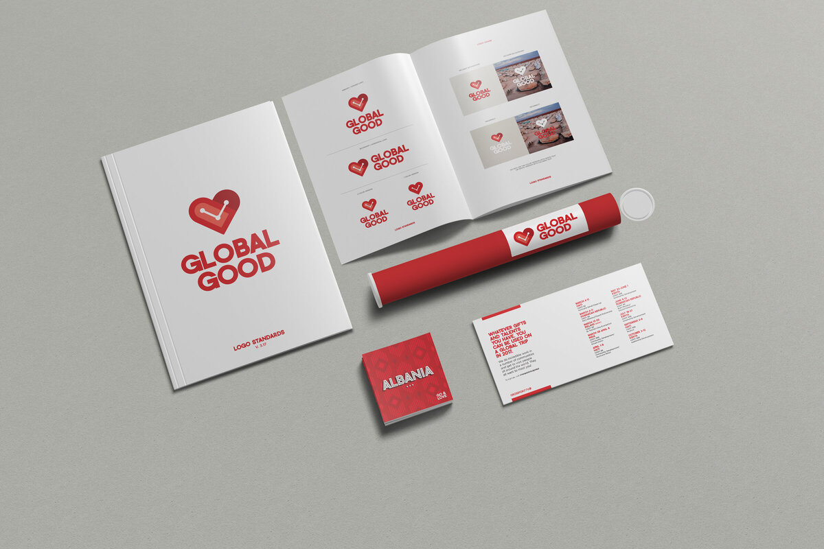 A flat lay of branded materials for 'Global Good,' including a logo standards booklet, an open brand guide with logo variations, a red mailing tube, a square booklet labeled 'Albania,' and a printed event or donation information card. The branding features a red heart with a graph line inside, paired with bold red text.
