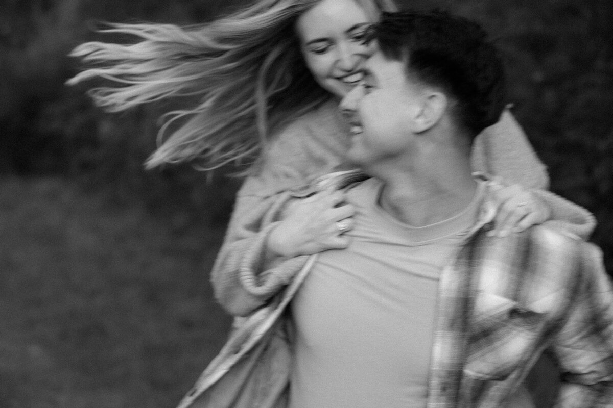 Man gives his fiance a piggy back ride as her hair blows in the wind during golden hour. 