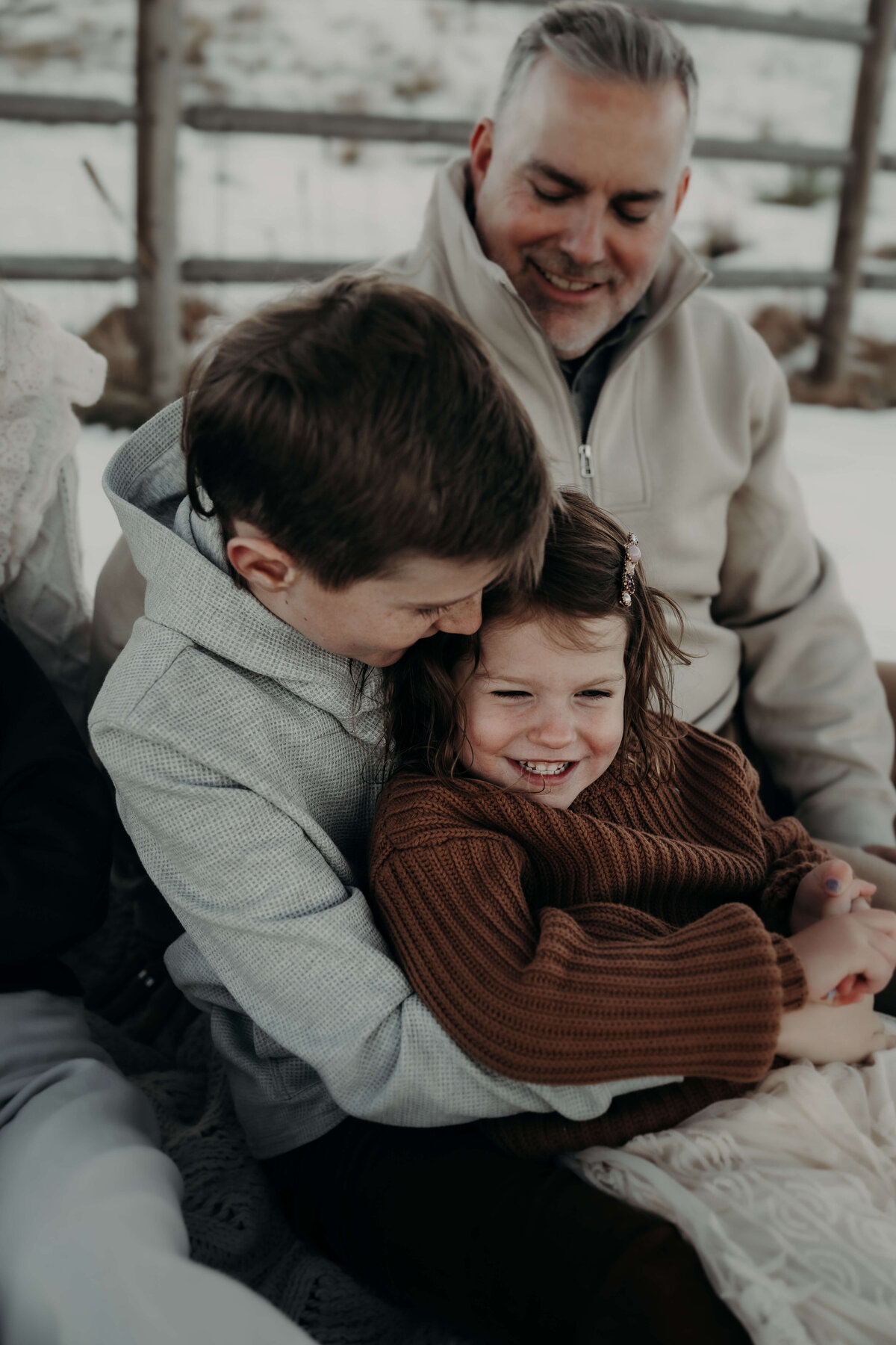 A big brother lets his little sister sit on his lap while he whispers something funny in her ear to make her laugh during a family photography session in Denver, Colorado.