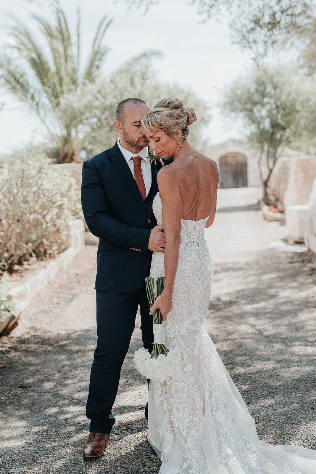 Bride looks gently down her shoulder holding white rose bouquet in lace wedding dress