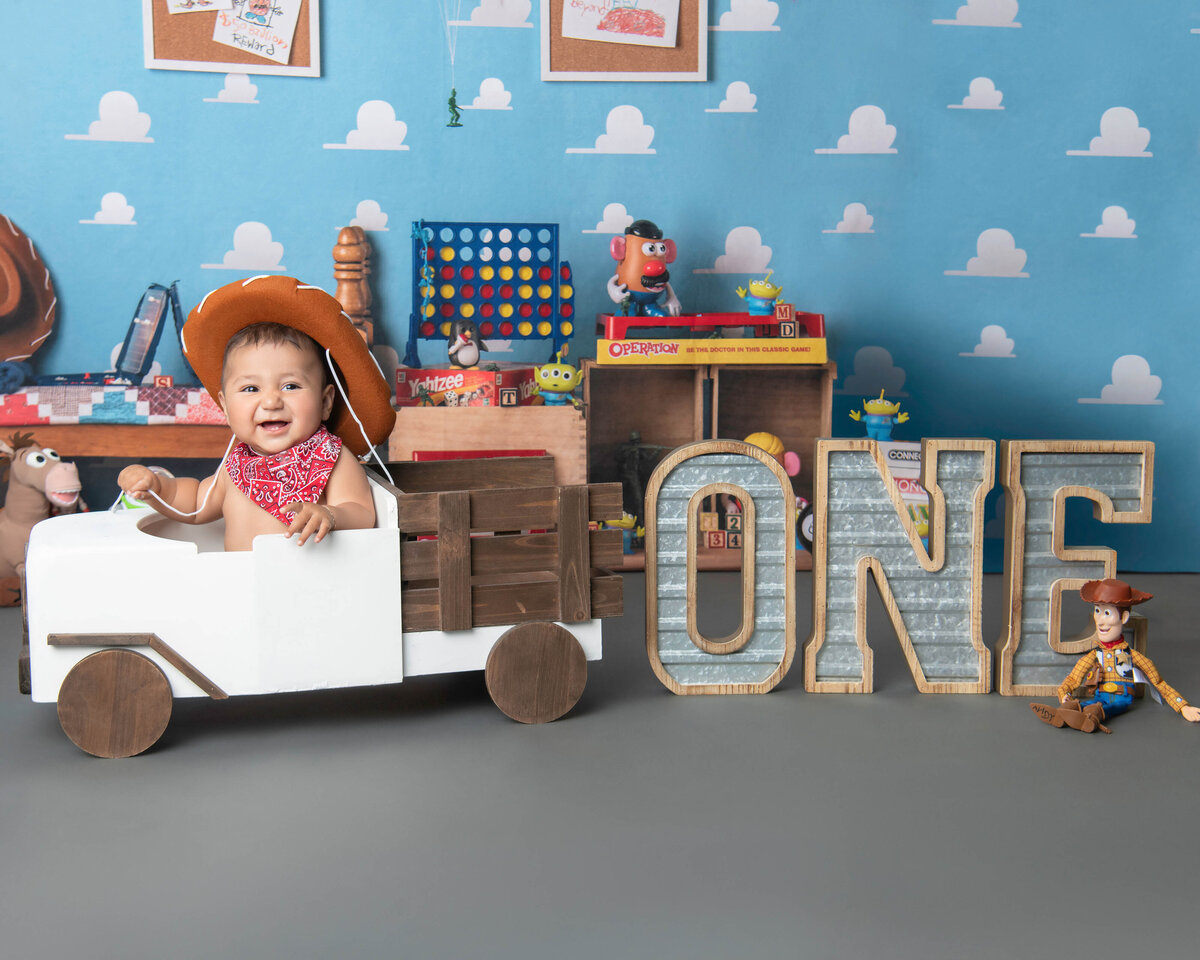 A giggling baby sits in a wooden truck dressed as a cowboy for his birthday in the studio of an Orange County milestone photography