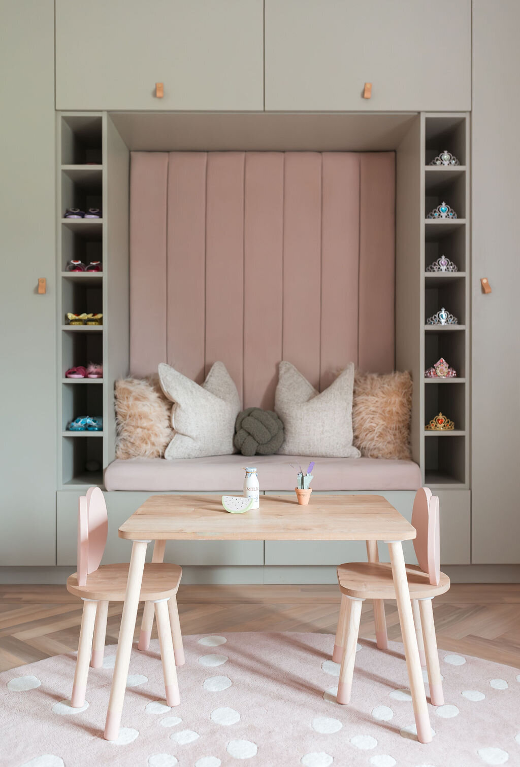 A cozy children's playroom with pastel-colored decor. It features a built-in cushioned seating nook with shelves and a wooden table with pink chairs.