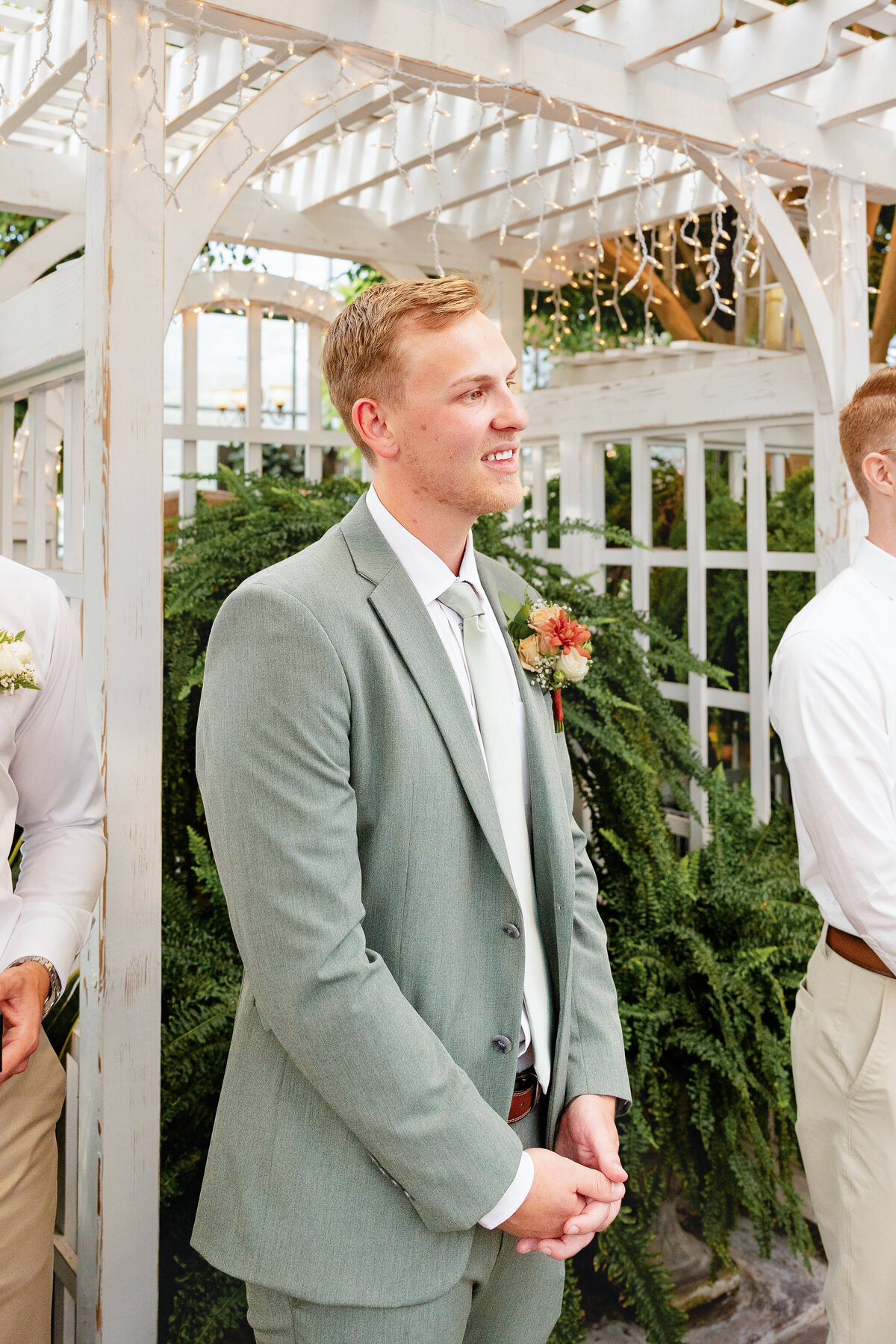 The groom in a sage green suit stands under a white pergola adorned with twinkling lights, smiling as he sees his bride for the first time.