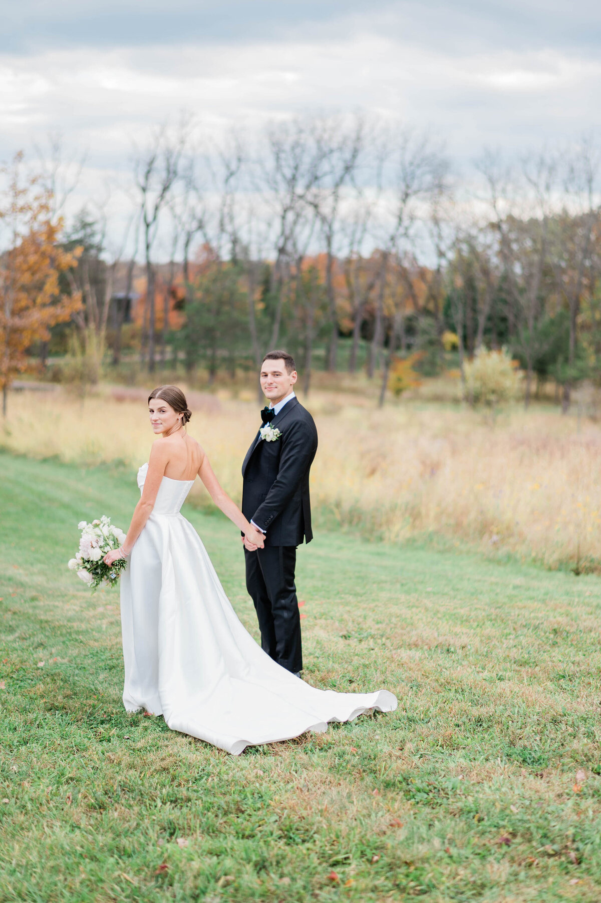 The couple holds hands in an open landscape, looking back at the camera