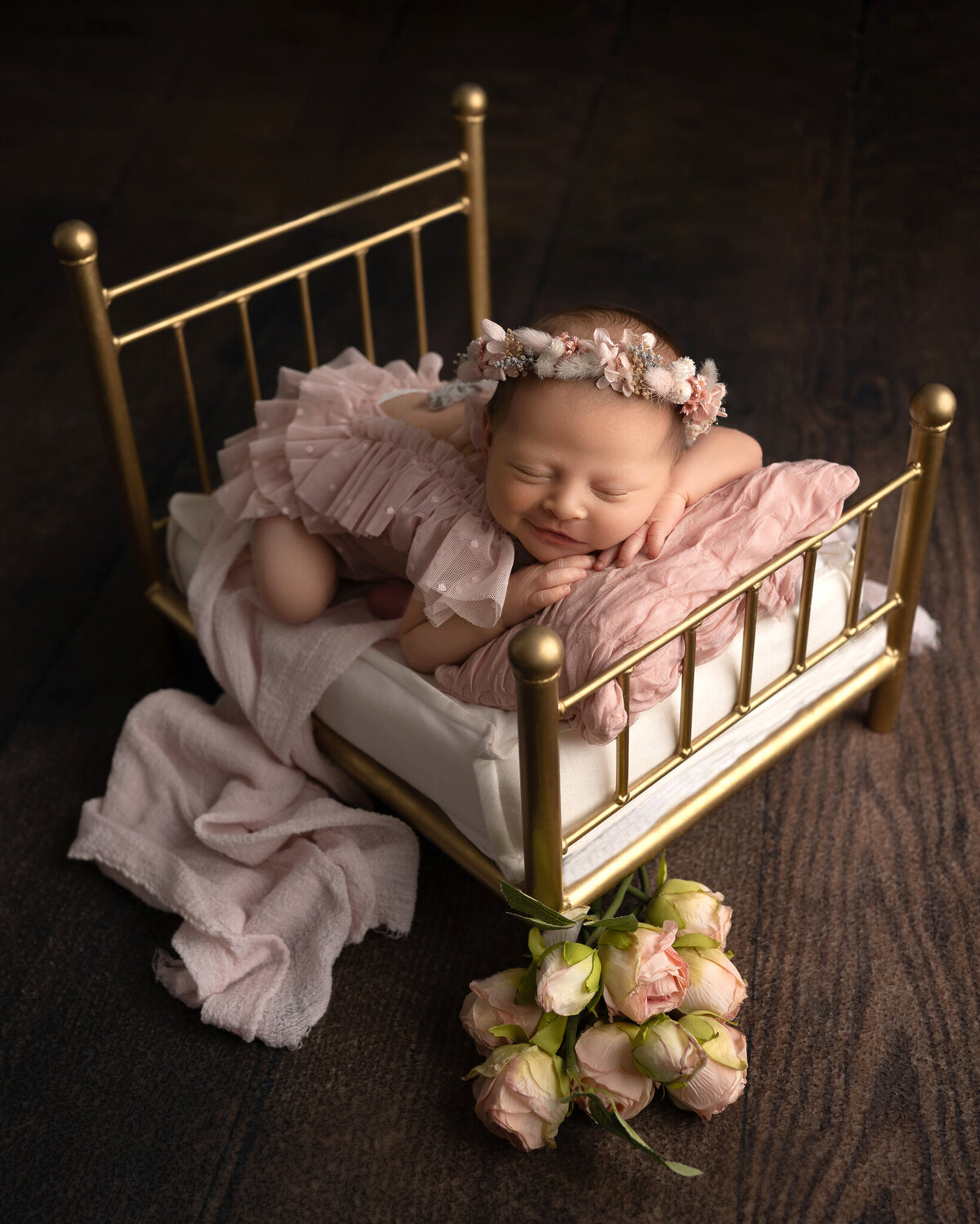Baby girl smiling in froggy pose on vintage gold bed with soft pink outfit and floral crown | High-end newborn studio in Lakeway and Austin, TX