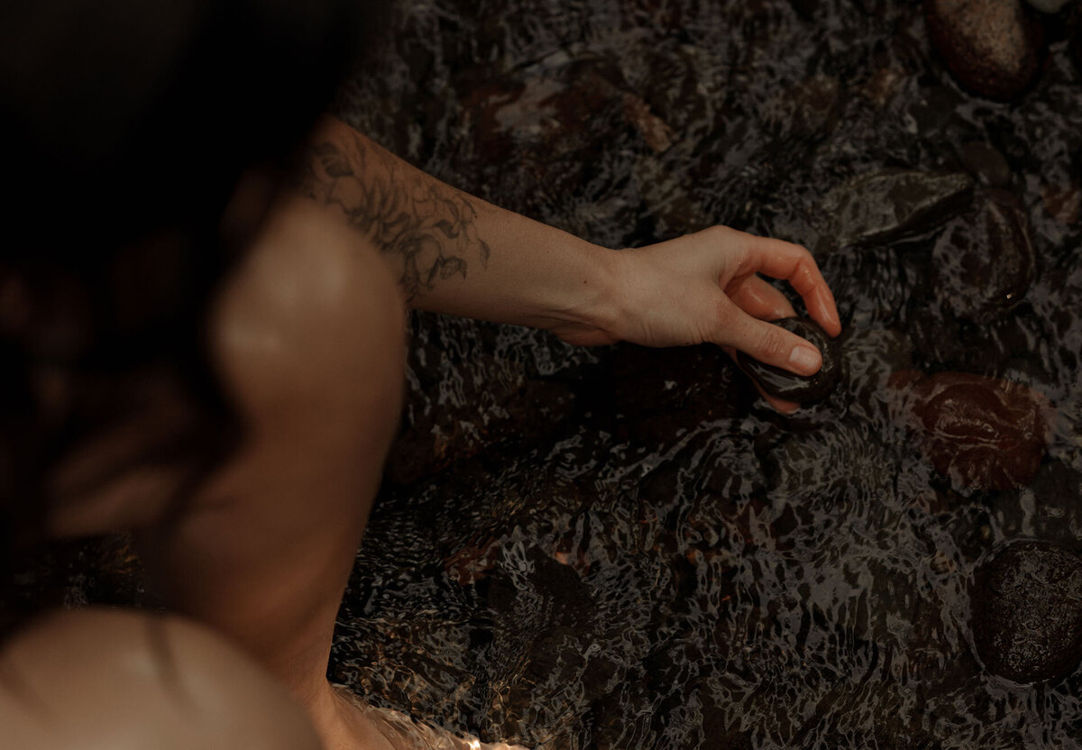 woman with tattoos sits near the river and observes river rocks in Bend, Oregon