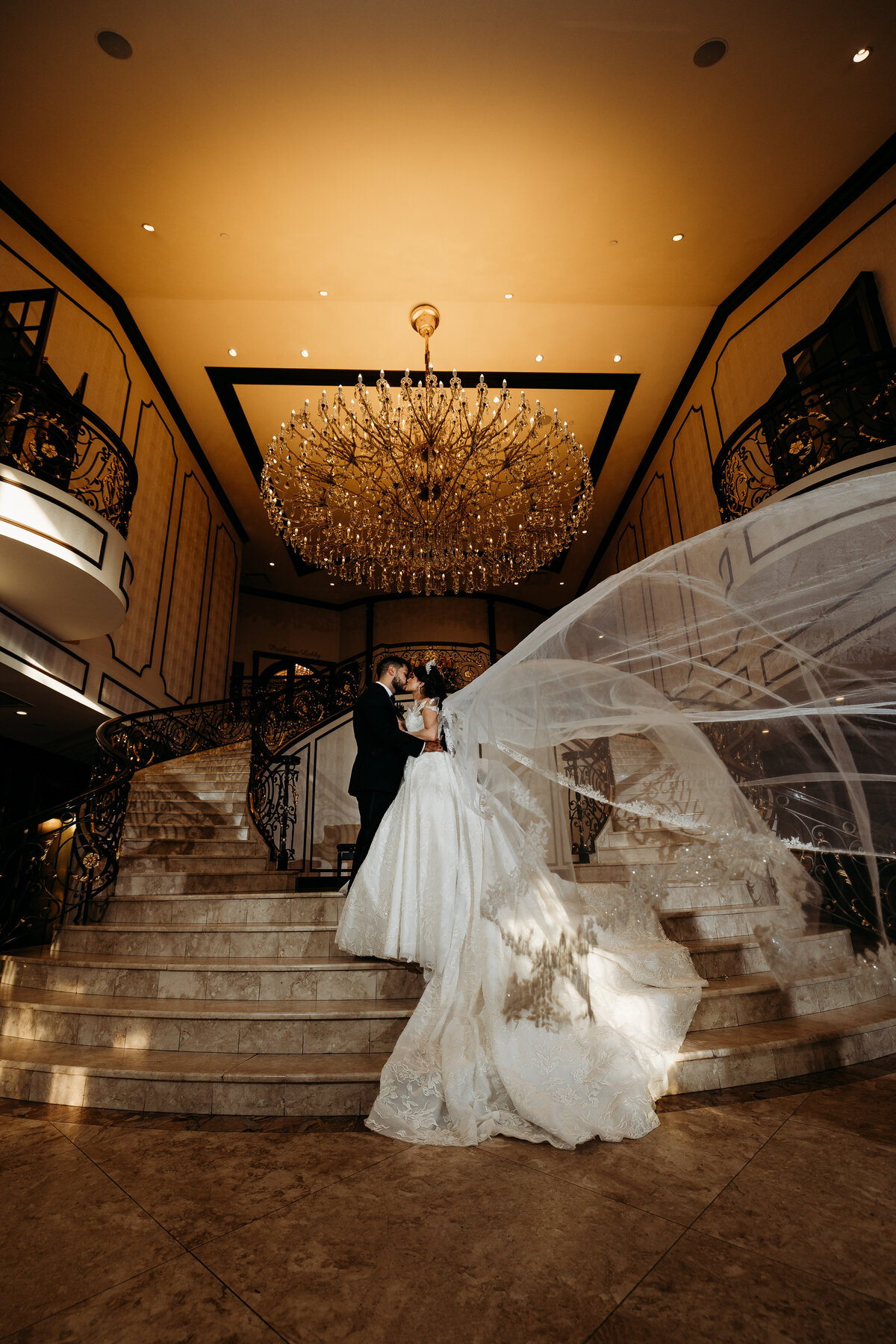 Bride and groom embrace on grand staircase as veil blows dramatically