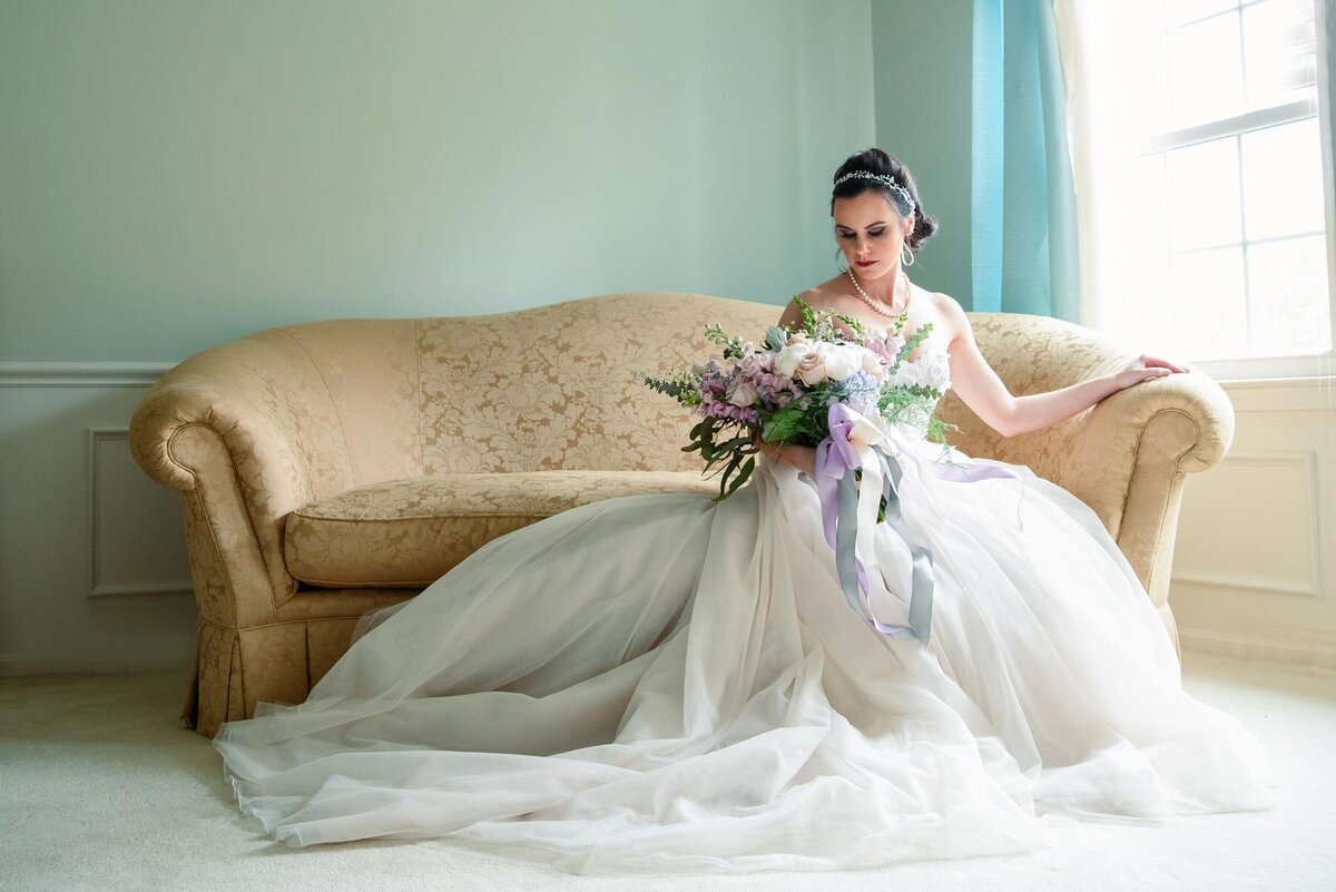 Bride sits elegantly on gold couch as dress spills off the couch and onto the floor around her, while she holds her bouquet
