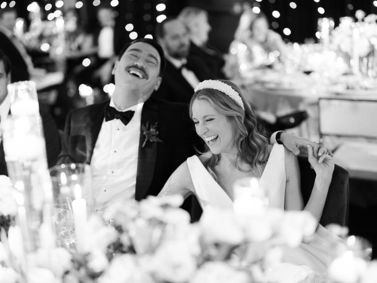 Black and white photo of a bride and groom laughing joyously at their wedding table, surrounded by guests.