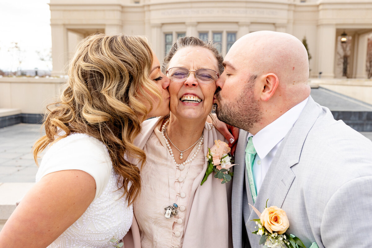 The bride and groom kiss an older woman on the cheeks, all smiling and laughing joyfully, captured in front of the Payson Utah LDS Temple.