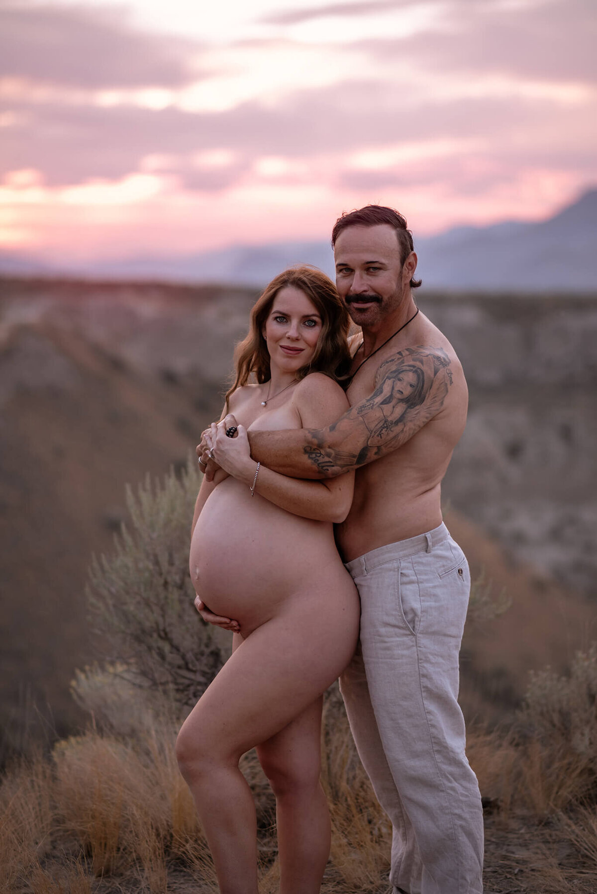 A husband and wife pose at blue hour in Kamloops for a stunning photo of them embracing. He holds her from behind while covering her breasts/ They both smile at the camera.