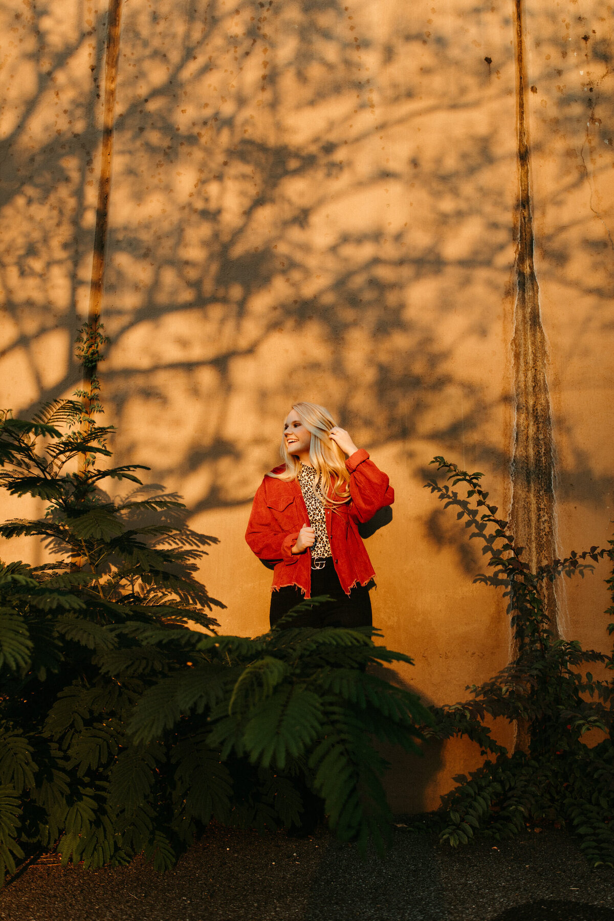 High school senior  in red jacket leaning against a wall downtown at sunset