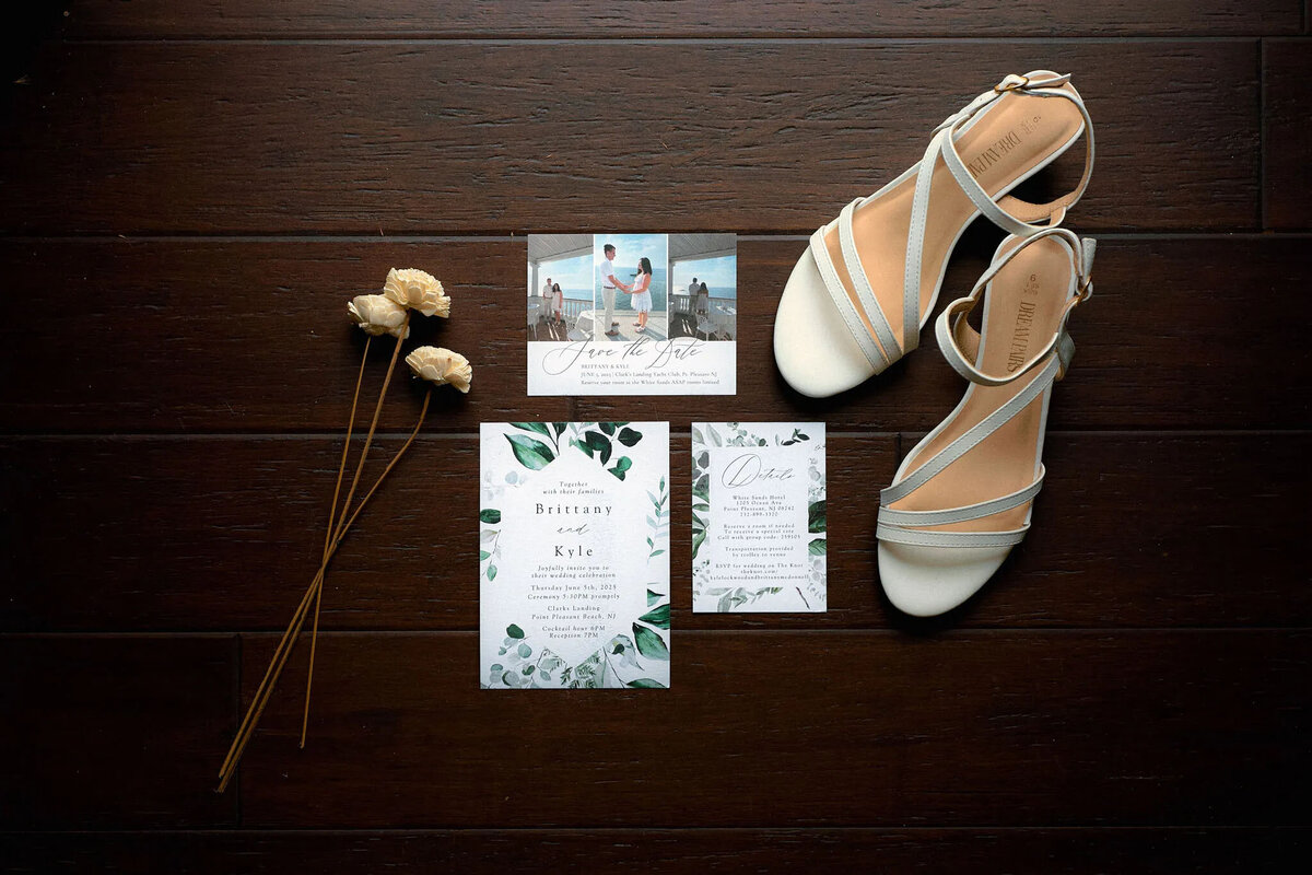 Flat lay of white heeled sandals, three dried flowers, and wedding invitations arranged on a dark wooden floor—captured by an NJ wedding photographer. The invitations feature floral designs and green leaves.