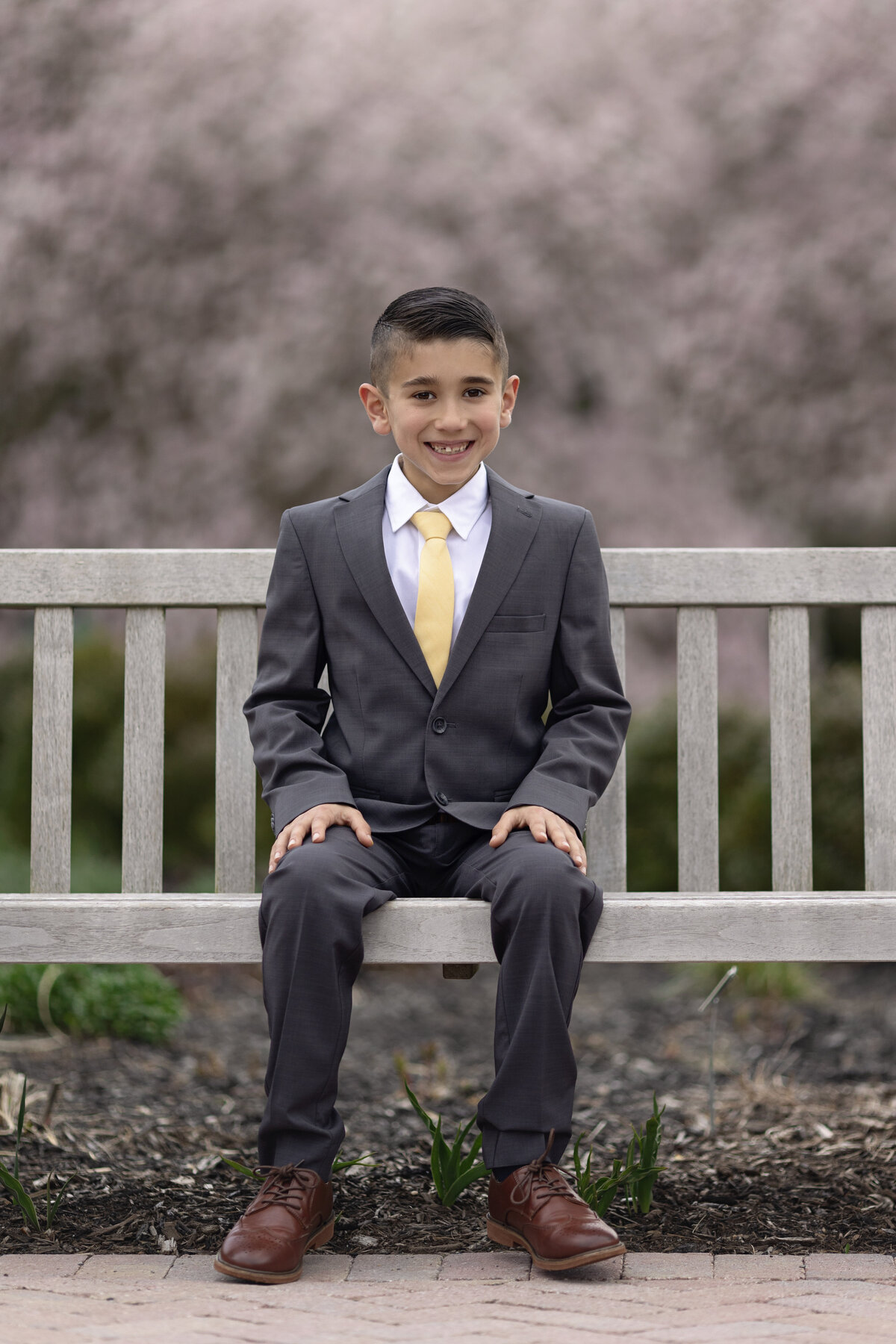 A young boy sits on a park bench in a grey suit and yellow tie smilingNew Jersey Communion Portrait Photographer