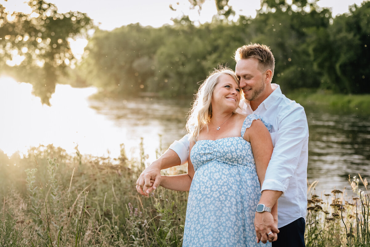 The couple stands by a river at sunset, with the man holding the woman from behind as they smile warmly at each other.