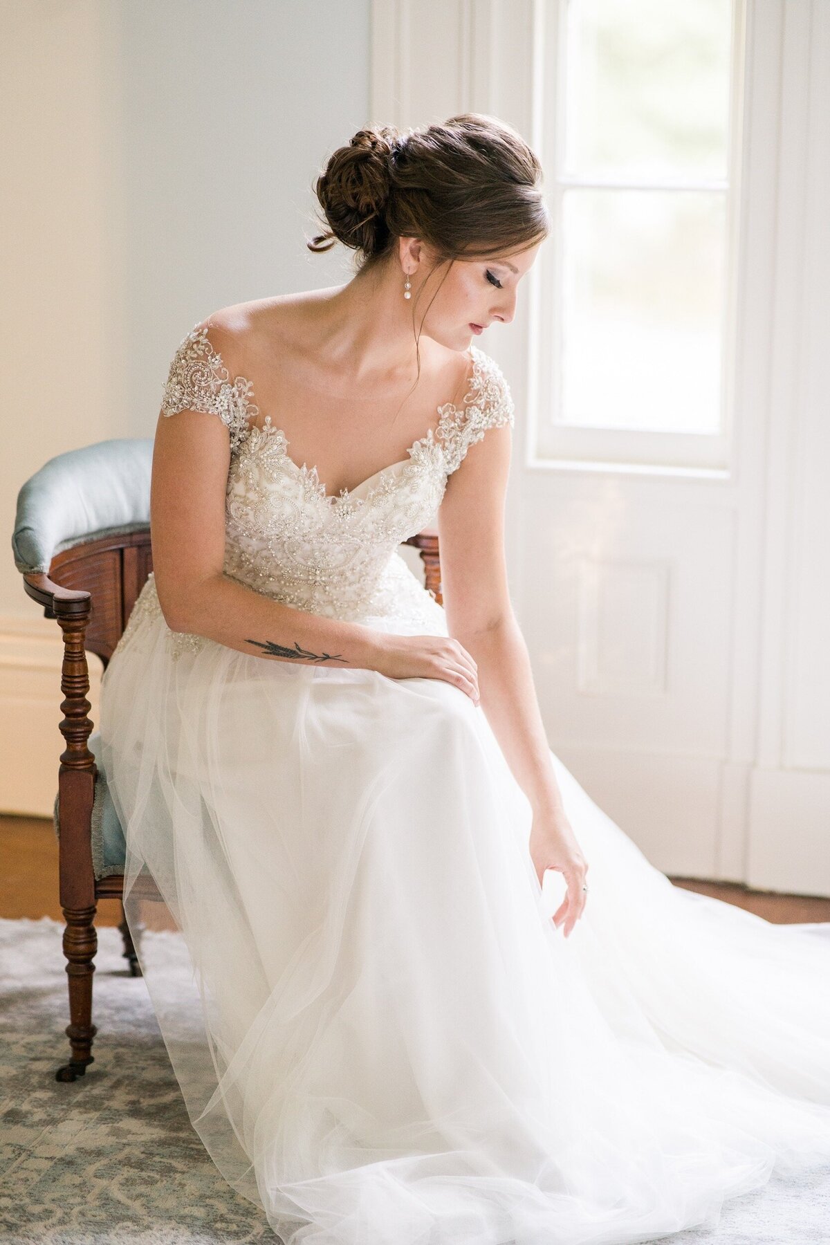 A bride reaches down to adjust her dress while sitting in a brightly lit bridal suite at Holt House in Lexington, NC near Charlotte, NC.