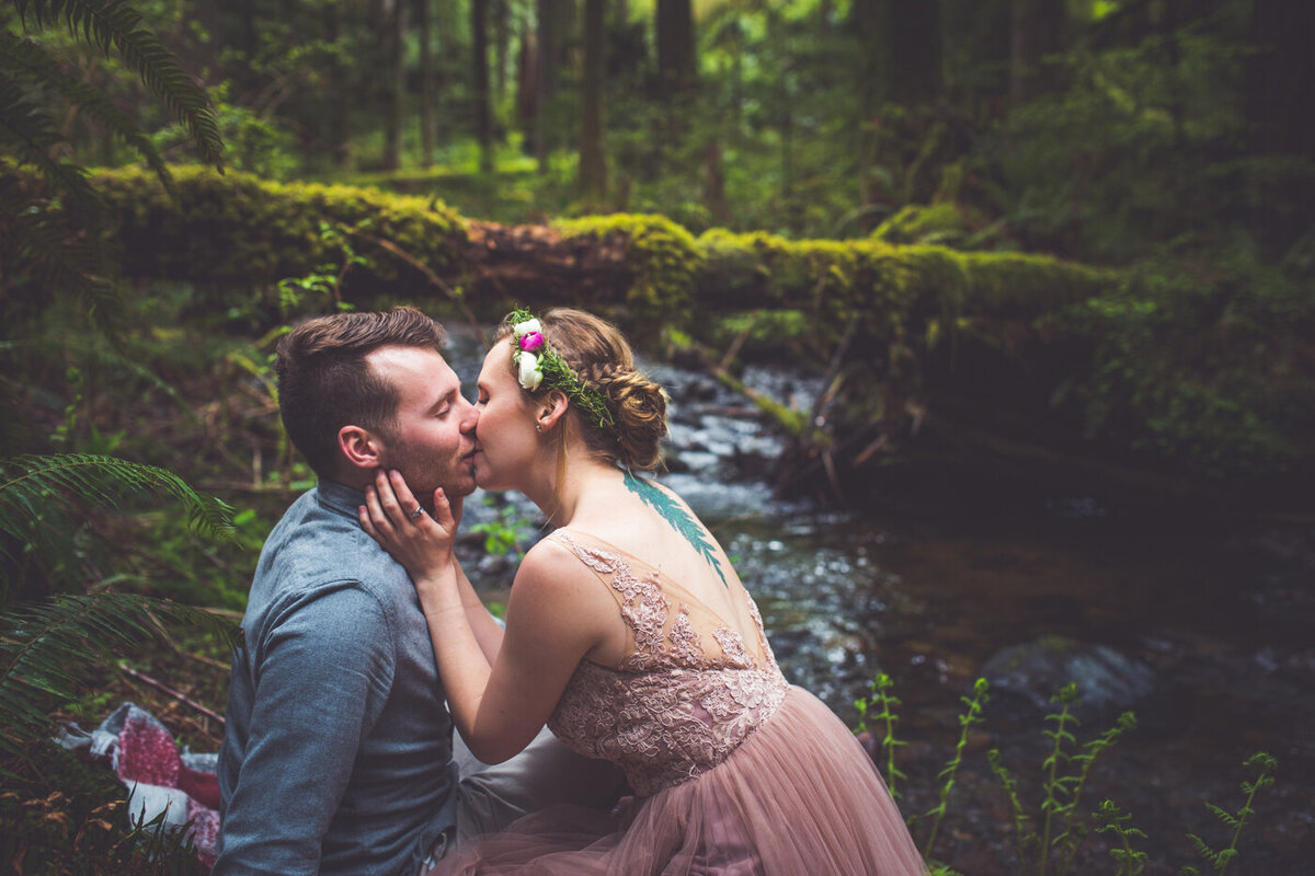 Whimsical and dreamy wedding photo of a bride and groom kissing by a creek in the forest.
