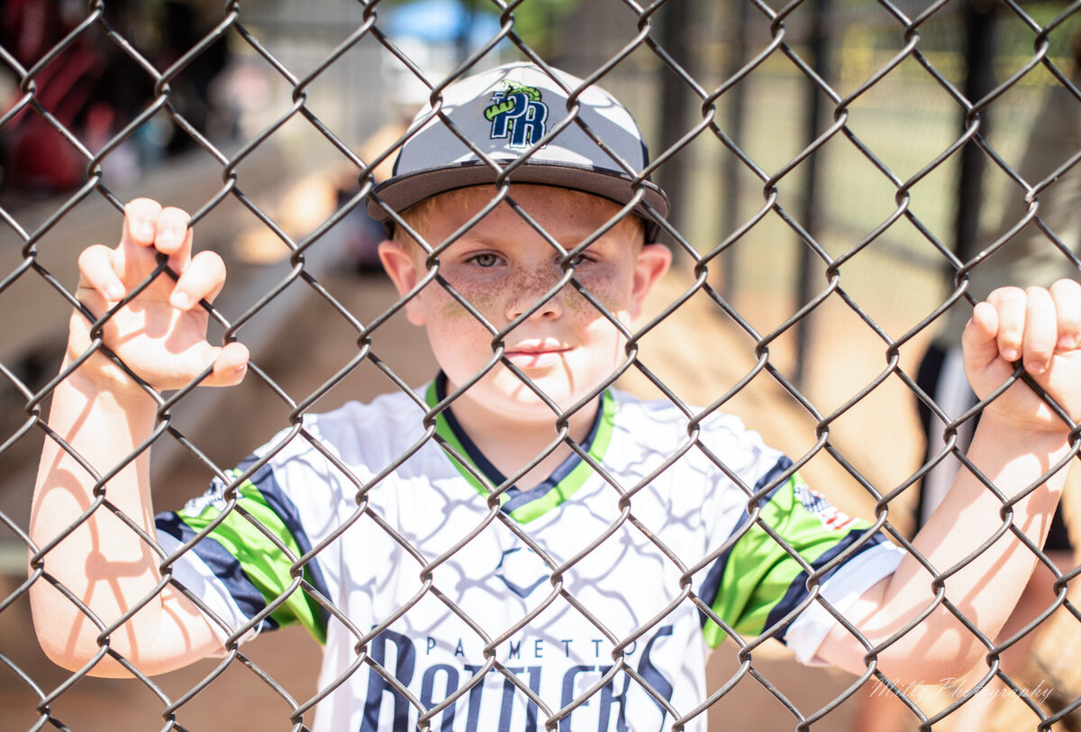 A small boy in a baseball uniform posing for his lifestyle photo behind a fence photographed by Millz Photography in Greenville, SC