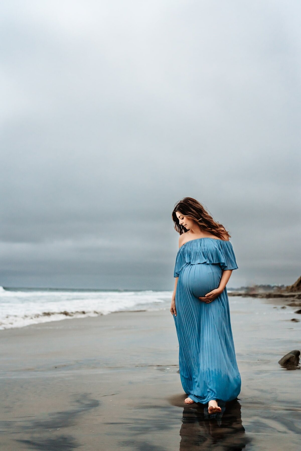 A pregnant woman in a long blue dress  holds her belly and walks on a Del Mar beach during a lifestyle maternity photo session by San Diego lifestyle maternity  photographer Love Michelle Photography