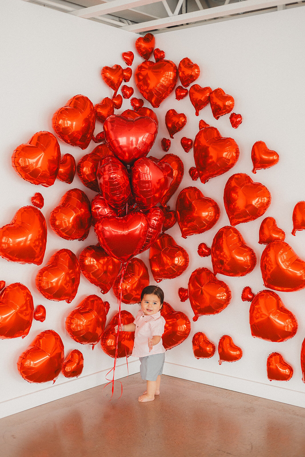 A baby boy stands in front of red heart balloons.