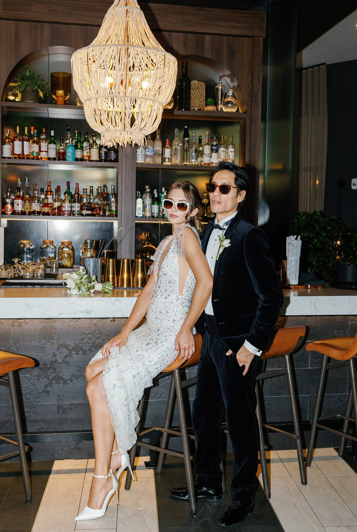 Bride and groom in stylish outfits posing at a bar with a chandelier, captured by Claudia Amalia, Miami wedding and lifestyle photographer.