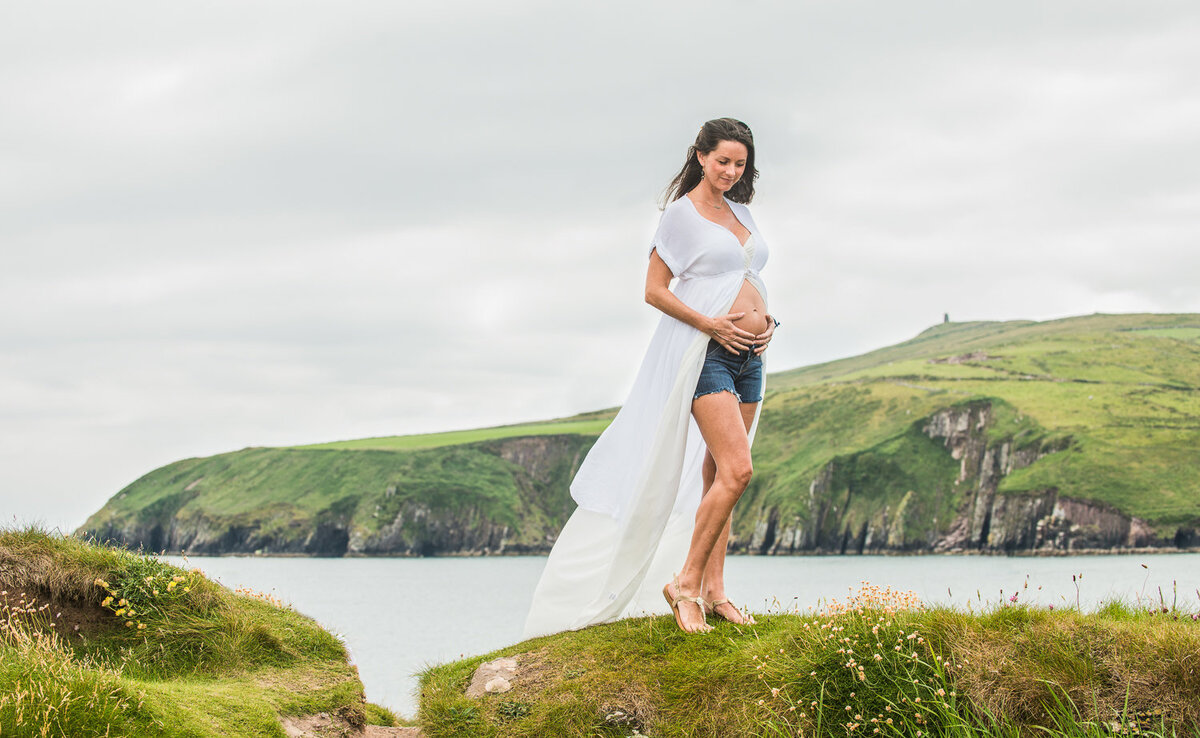 portrait of pregnant woman with long black hair, wearing a flowing white dress standing on a cliff