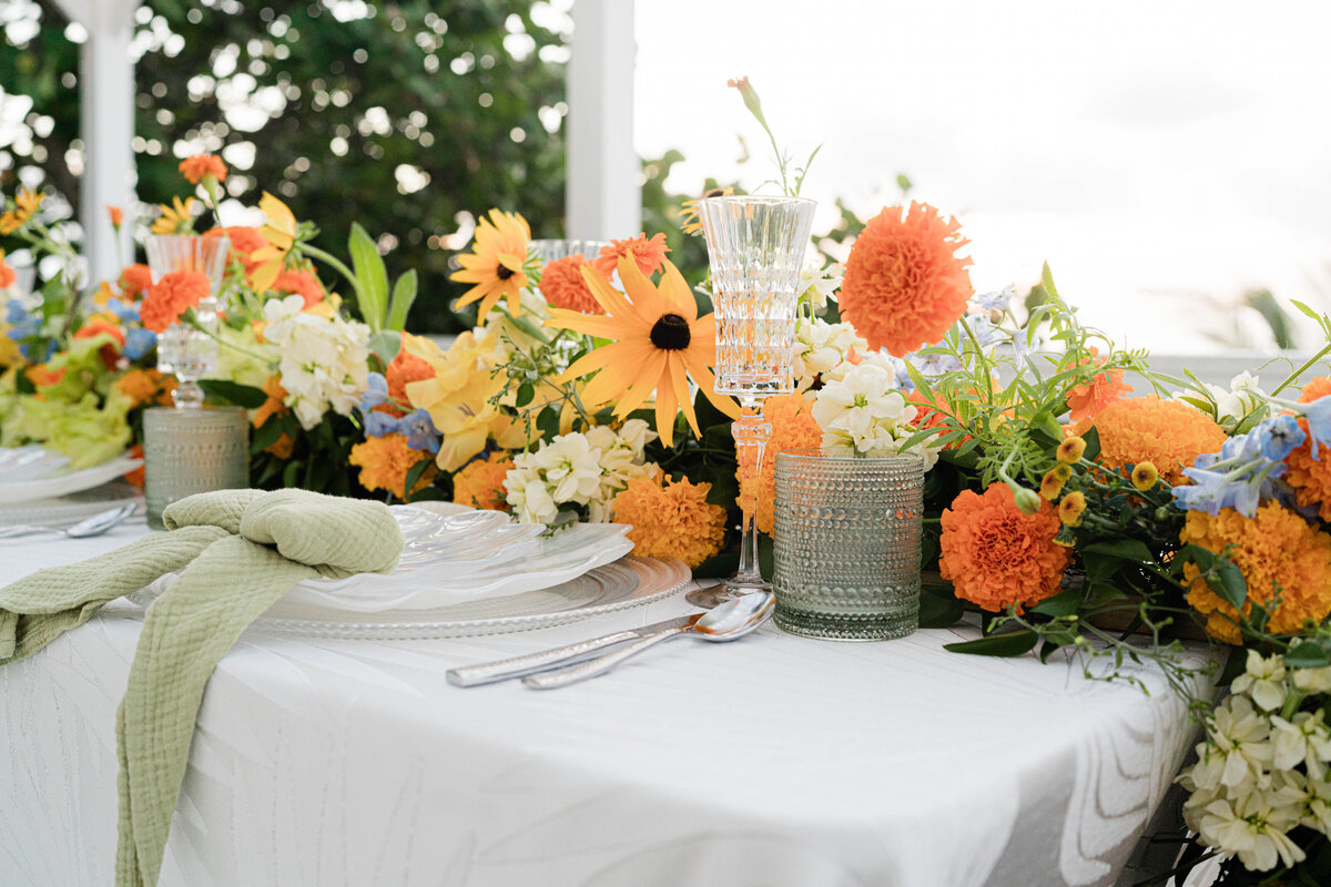 white on white palm leaf tablecloth with hammered silver flatware and an iridescent seashell plate adorned with a white, orange, blue and green floral table runner