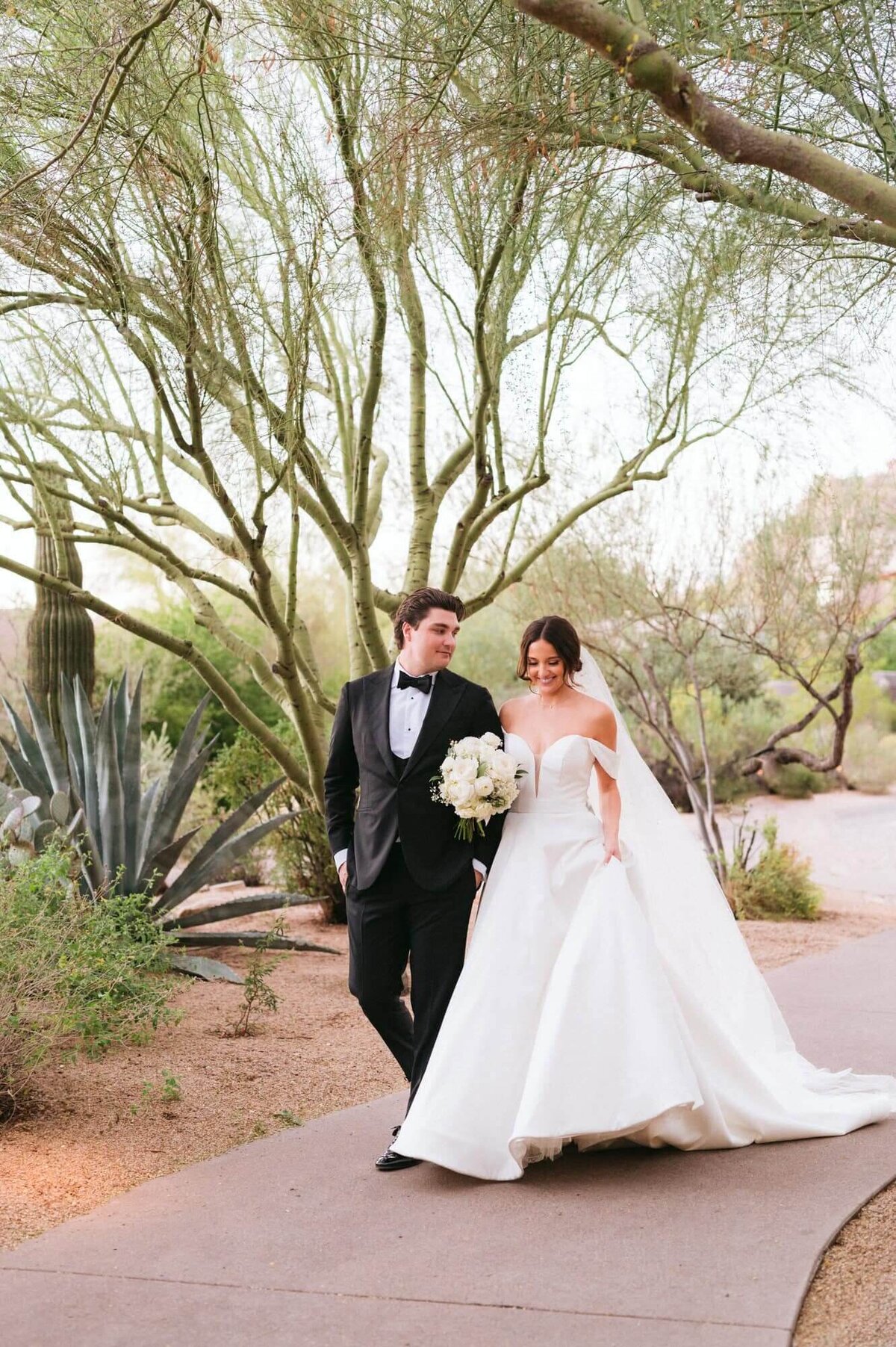 Bride and groom walking together through the desert pathways at Four Seasons Troon North in Scottsdale, surrounded by cacti and desert trees.