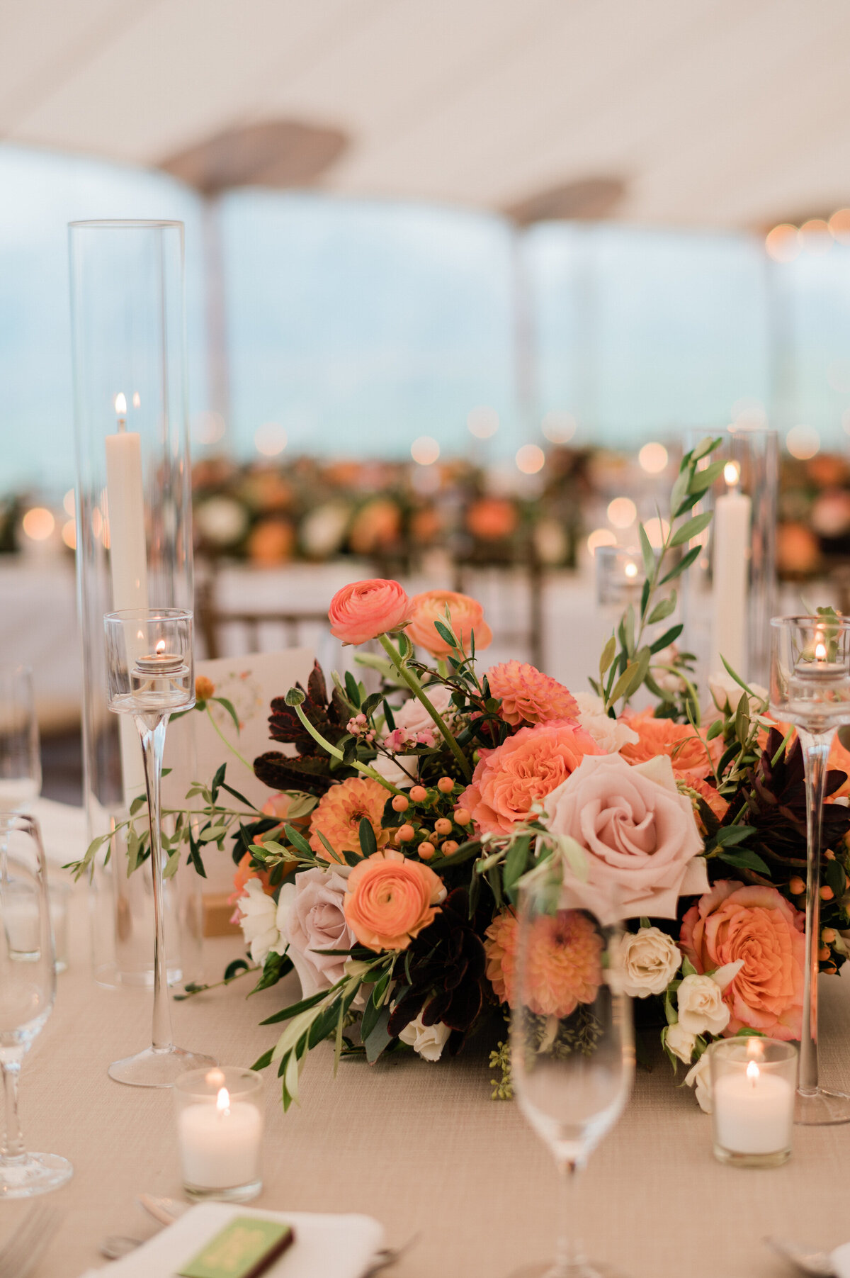 A floral centerpiece with peach and blush roses and greenery sits atop a linen-covered reception table.
