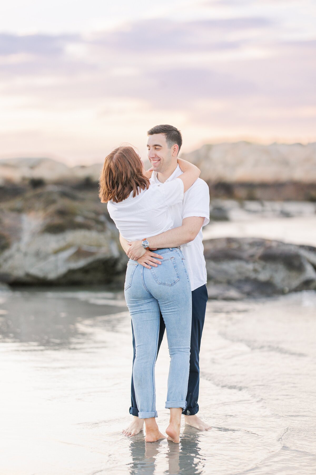 A man and woman hugging on Wingaersheek Beach with the rock and sunset in the background during their engagement photography session.