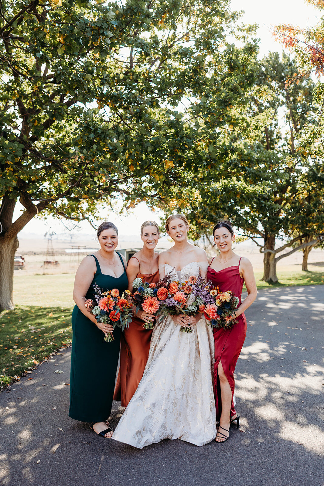 Bride and her bridesmaid in mismatching colours holding their bouquets. They are standing under trees at Petrichor Farm, a rural wedding venue near Canberra