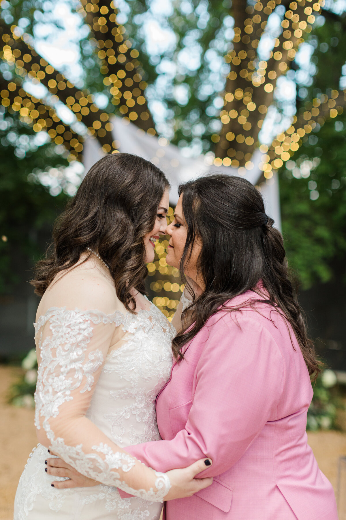 Portrait of two brides holding each other close and coming in for a kiss on their wedding day at Artspace111 in Fort Worth, Texas. The bride on the left is wearing a long sleeve, intricate, white dress while the bride on the right is wearing a pink suit. A large tree covered in lights towers above them in the background.