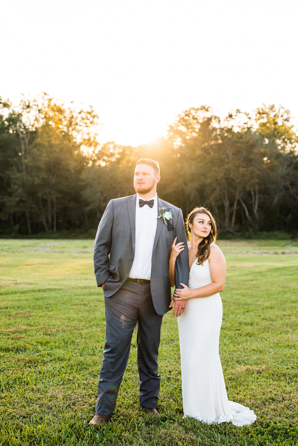 bridal portrait by Virginia wedding photographer with bride holding grooms arm and looking to the distance while the sun sets over the tree line in the distance