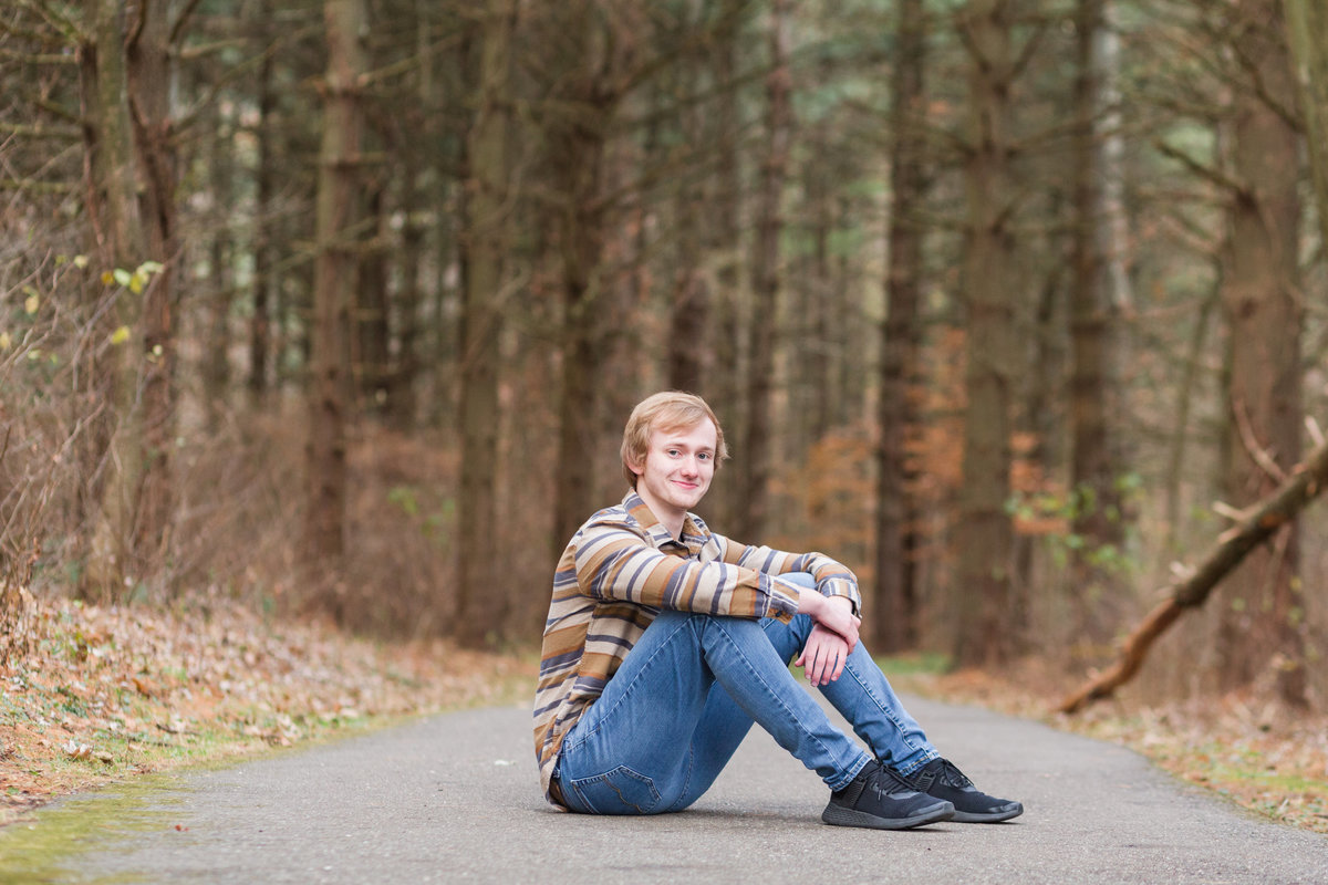 saint thomas equainus senrior guy sitting on paved walkway with tall pines in the background, photographed at quail hollow in hartville, photographed by Jamie Lynette Photography canton ohio senior photographer