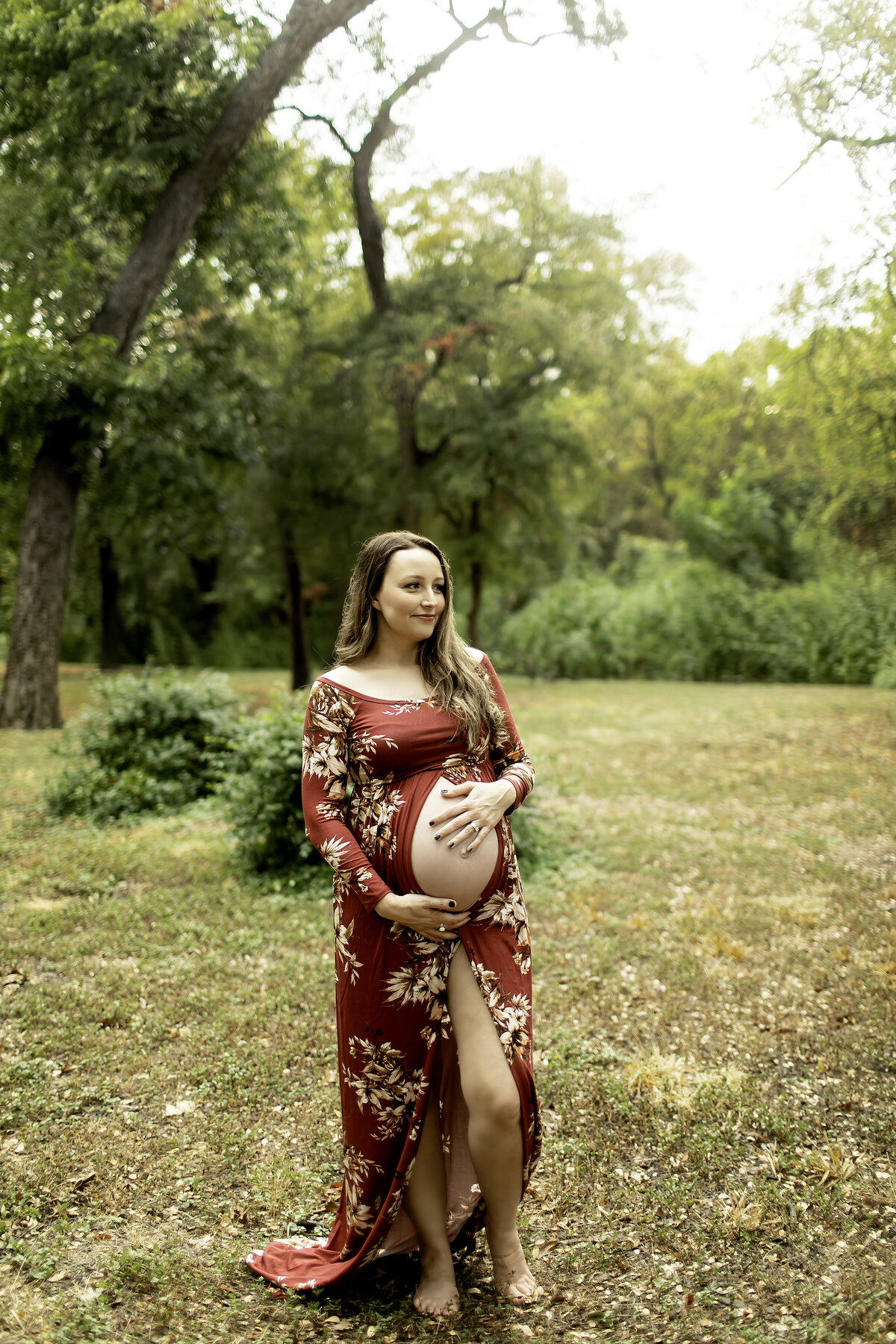 A serene maternity moment of the mom-to-be wearing a red dress, gazing lovingly at her belly while surrounded by the scenic beauty of Trinity Park, captured by Chunky Monkey Photography.