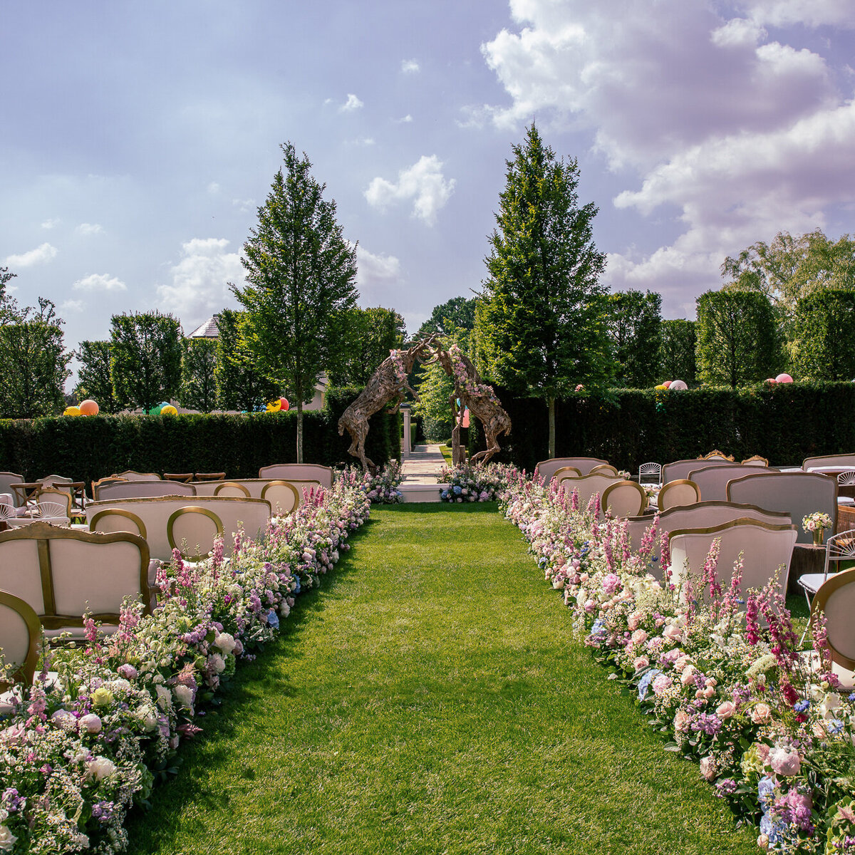 A luxurious wedding ceremony aisle is filled with planted flowers, and mis matched seating in a gorgeous garden.