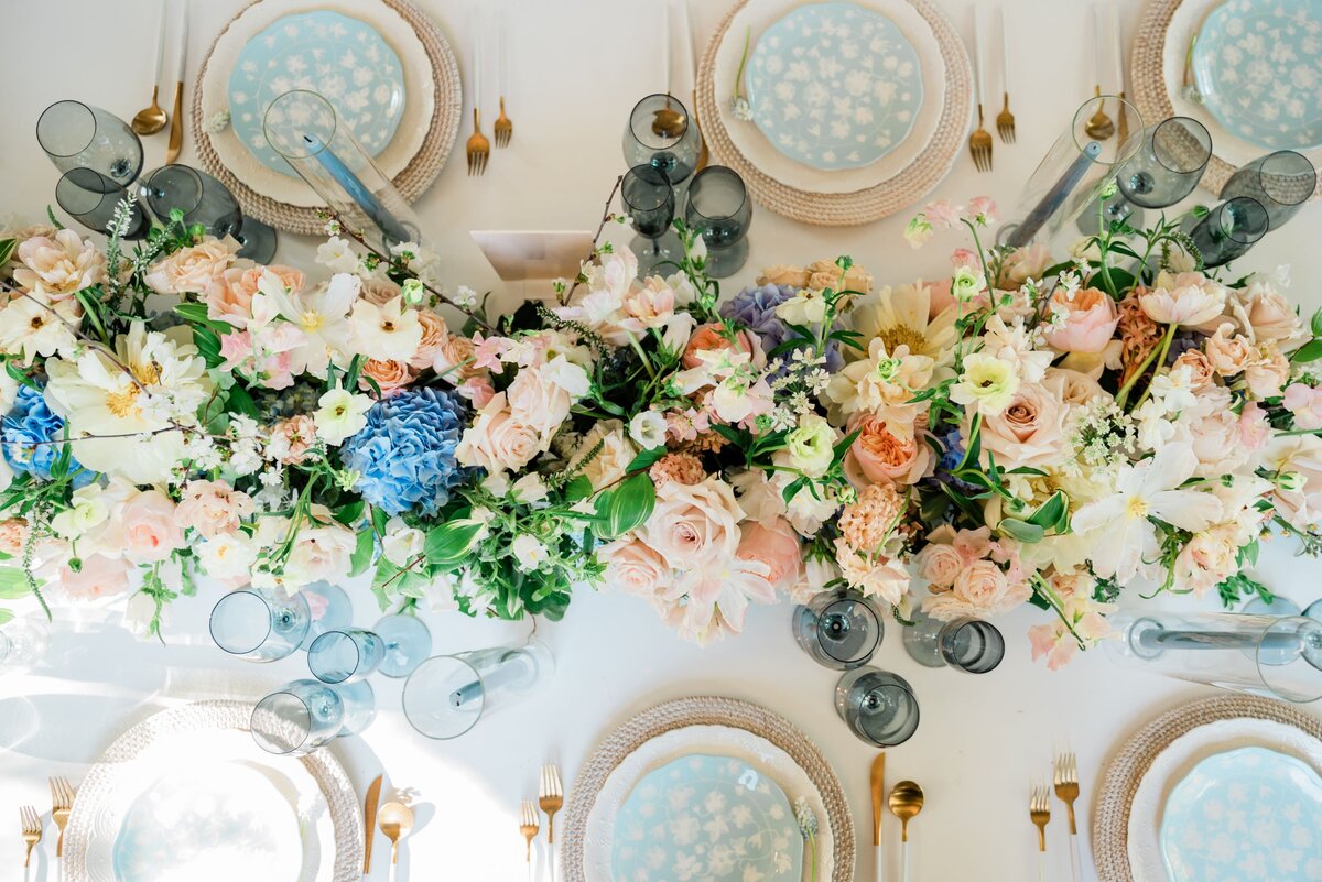 A flatlay of a wedding guest table with blue, pink, and yellow flowers as the centerpiece