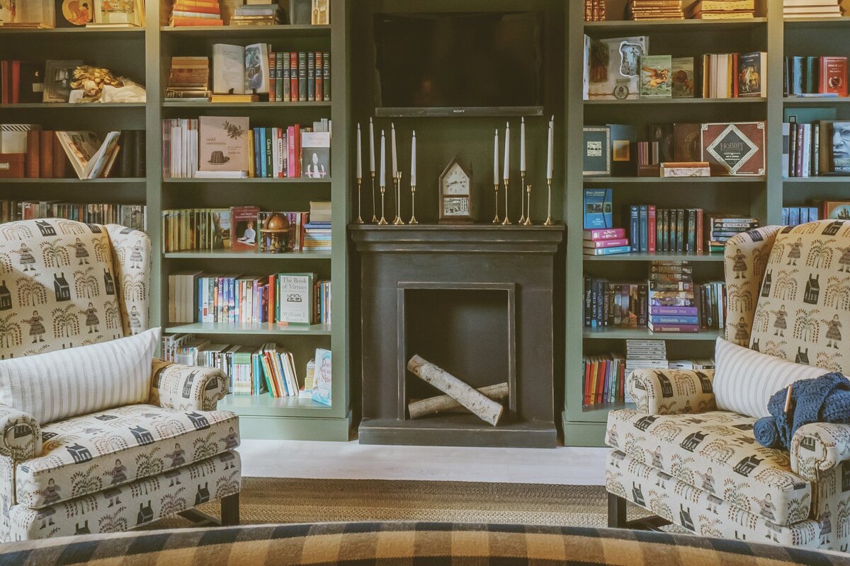Green built-ins library shelves filled with books and faux fireplace with birch logs and candlesticks stop the fireplace, woven rug in warm mocha tones and two early American textile armchairs pair