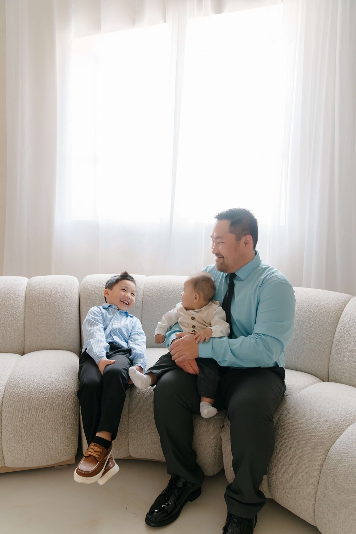 Father sharing a light-hearted moment with his two sons on a plush studio couch with natural window light.