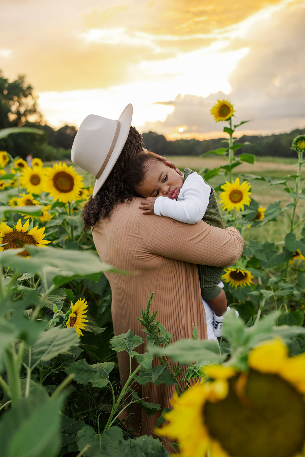Mom holds infant son as he rests his head on mom's shoulder in sunflower field