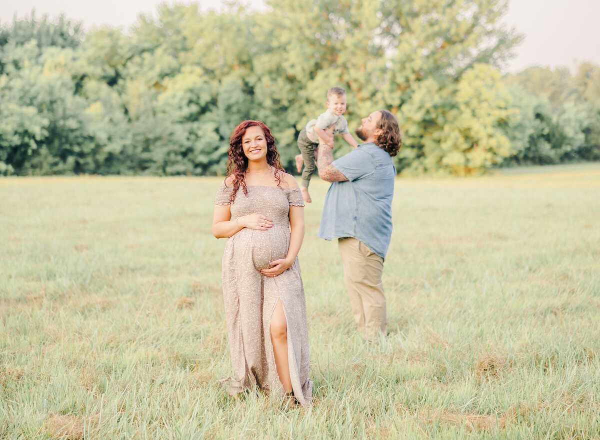 Mom standing in field showing pregnant belly with dad holding son in the distance
